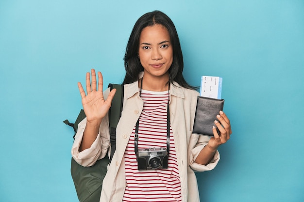Filipina mit Kameratickets-Rucksack auf blauem, fröhlichem Lächeln zeigt Nummer fünf mit den Fingern