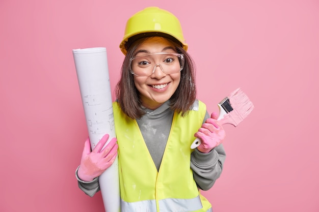 Filmación en interiores de alegre constructora ocupada con la renovación de la casa sostiene el pincel y el plano del edificio viste uniforme aislado en rosa