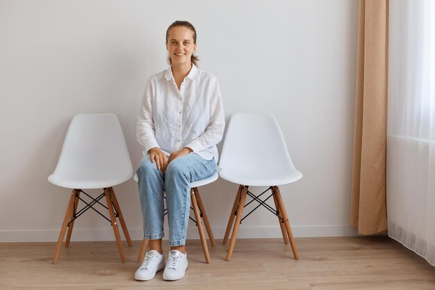 Filmación en interiores de una atractiva mujer adulta joven sentada en una silla contra la pared blanca, vestida con camisa y jeans, posando con una sonrisa, mirando felizmente a la cámara, expresando optimismo.