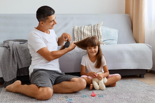 Filmación en interiores de un atractivo hombre vestido con camiseta blanca sentado en el suelo y peinando a su linda hijita, cuidando a su hija, pasando un rato agradable juntos.