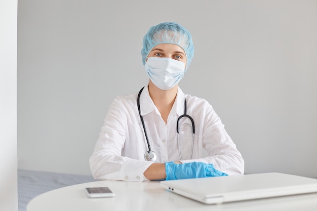 Filmación en interiores de una doctora seria y segura con gorro médico, mascarilla quirúrgica, guantes de goma y una bata, usando el portátil para trabajar, mirando a la cámara mientras está sentado en la mesa.