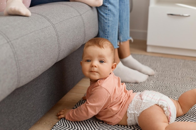 Filmación en interiores de feliz lindo bebé adorable niña arrastrándose por el piso en el piso de la sala de estar, mirando a la cámara con ojos curiosos, vistiendo camisa y pañal.