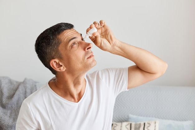 Foto filmación en interiores de un hombre caucásico moreno aplicando gotas para los ojos que tienen problemas con el tratamiento de la vista.