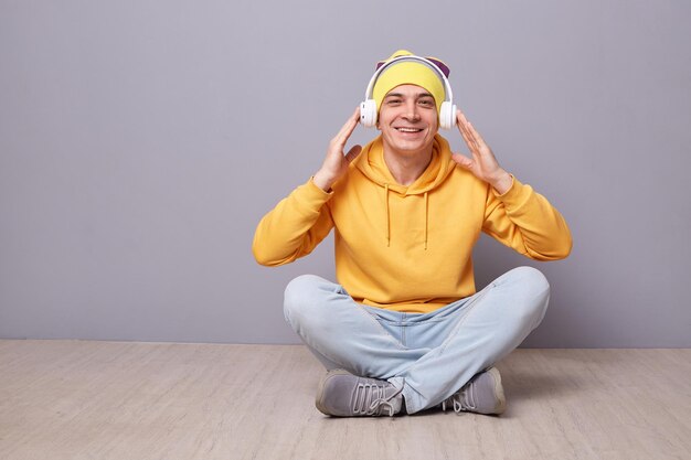 Filmación en interiores de un joven adulto caucásico sonriente y encantado con ropa informal sentado en el suelo contra una pared gris mirando a la cámara escuchando música