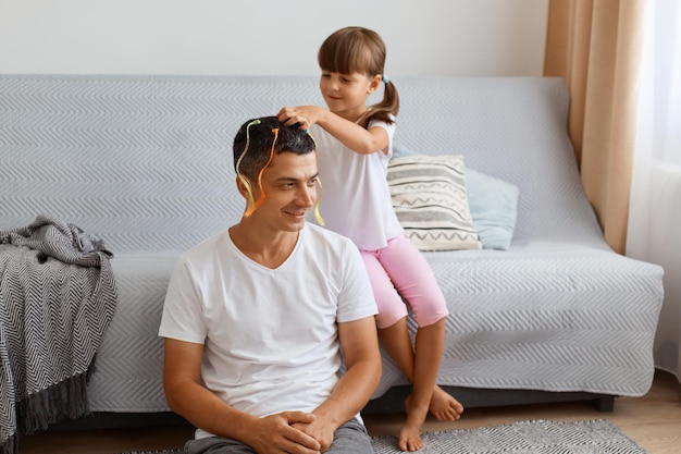 Filmación en interiores de un joven en casa con su pequeña niña linda, hija haciendo peinado para el padre, pasando tiempo juntos, sentados en el piso cerca del sofá en la sala de estar.