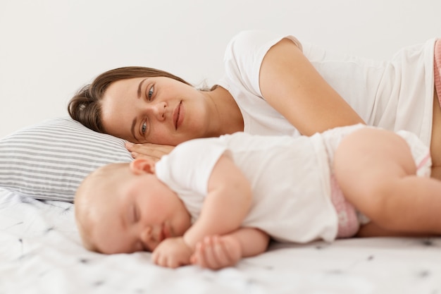 Filmación en interiores de una joven madre adulta acostada cerca de su bebé durmiendo, mirando a la pequeña hija con gran amor, mujer vestida con camiseta blanca de estilo casual, posando en casa por la mañana.