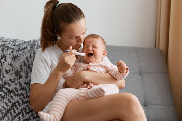 Filmación en interiores de la madre midiendo la temperatura de su bebé enfermo que llora sentado en el sofá de la sala de estar, un niño pequeño que se resfría, tiene gripe o gripe.