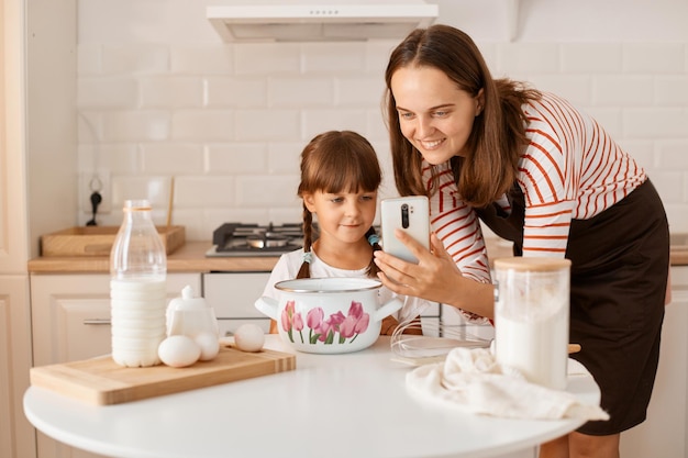 Filmación en interiores de una mujer adulta joven caucásica cocinando con su hija en la cocina, navegando por Internet para encontrar recetas, mirando sonriendo el dispositivo, felices de hornear juntos.