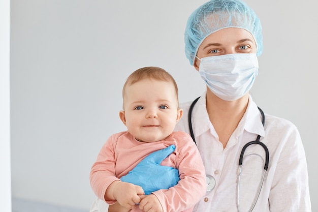 Filmación en interiores de una mujer médico de familia con uniforme médico y mascarilla quirúrgica sosteniendo al bebé en las manos, mirando a la cámara, examina la salud de los niños.