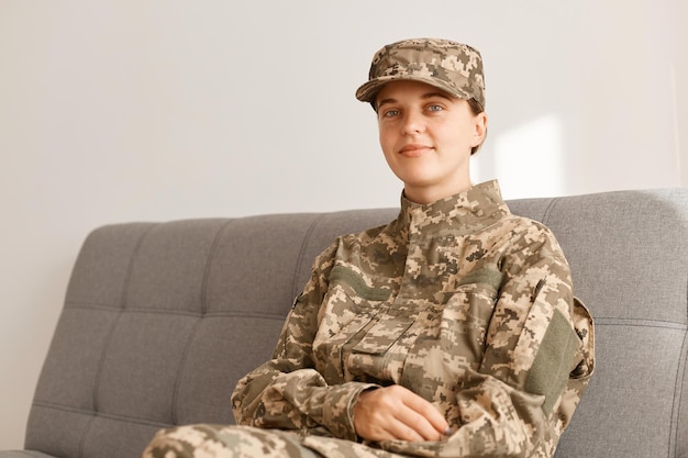 Filmación en interiores de una mujer militar positiva sonriente con uniforme de camuflaje y gorra, sentada en un cómodo sofá gris, mirando a la cámara con expresión facial optimista.