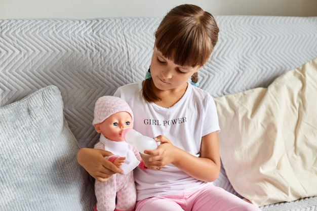 Filmación en interiores de una niña sentada en el sofá de la habitación en casa jugando con la muñeca, como mamá, niña en edad preescolar de cabello oscuro sosteniendo un juguete.