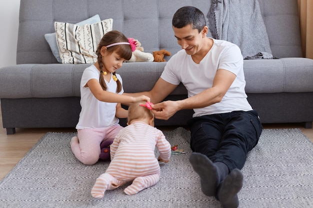 Filmación en interiores de un padre caucásico guapo morena jugando con sus hijas mientras está sentado en el piso cerca del sofá, vistiendo pantalones y camiseta blanca, expresando felicidad.