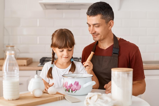 Filmación en interiores del padre de familia feliz y su linda hijita sentados a la mesa en la cocina y haciendo pasteles, rodeados de ingredientes para hornear galletas o pasteles.