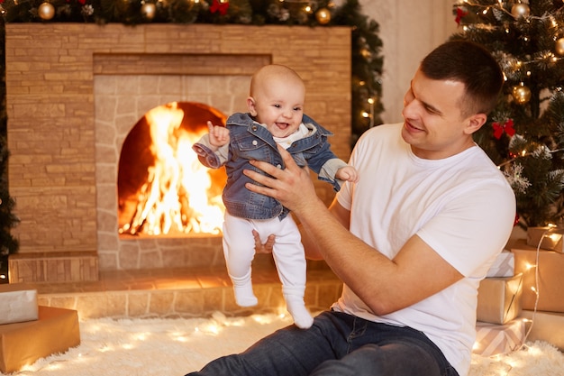 Filmación en interiores de un padre guapo con una camiseta blanca casual con una pequeña hija en las manos mientras está sentado junto a la chimenea y el árbol de Navidad en casa, feliz año nuevo.