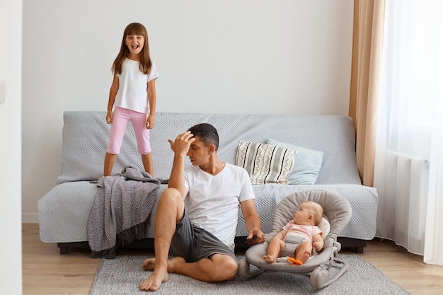 Filmación en interiores de serio y cansado padre moreno guapo, vestido con camiseta blanca y pantalones cortos, hace un comentario a su hija mayor, se porta mal, salta en el sofá.