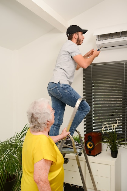 Filtro de aire de limpieza de electricista joven guapo en una unidad interior del sistema de aire acondicionado en una casa de cliente con una mujer mayor