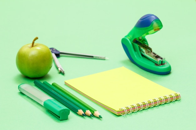 Filzstift, Farbstifte, Notizbuch, Apfel, Kompass und Hefter auf grünem Hintergrund. Zurück zum Schulkonzept. Schulbedarf. Geringe Schärfentiefe.
