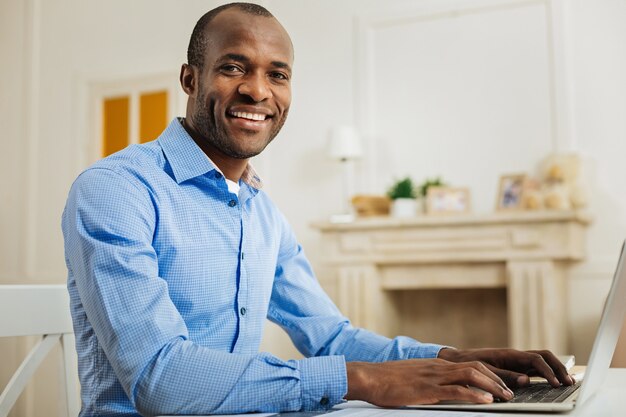 Fim de semana de trabalho. Homem afro-americano bonito e inspirado, sorrindo e trabalhando no laptop enquanto está sentado à mesa