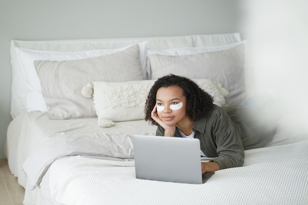 Fin de semana en la cama Hermosa chica afro aplica parches en los ojos y se relaja en casa viendo una película