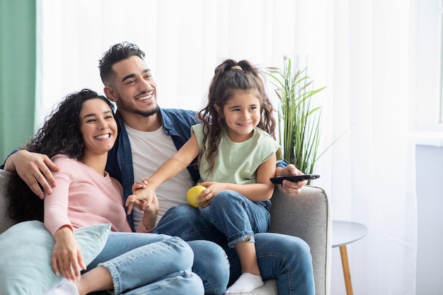 Fin de semana familiar. Alegres padres árabes y su pequeña hija viendo la televisión en casa, feliz esposo, esposa y niña del Medio Oriente relajándose en el sofá en la sala de estar, disfrutando del pasatiempo doméstico