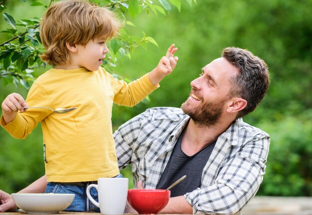 Fin de semana familiar Padre hijo come comida y diviértete Menú para niños Comida casera Hábitos alimentarios Niño pequeño con papá comiendo comida patio de picnic naturaleza fondo Desayuno de verano Concepto de comida saludable