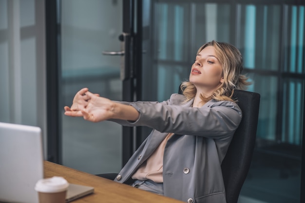 Finalización del trabajo. Mujer de negocios joven cansada que estira sus brazos cerrados hacia adelante mientras está sentada detrás de la computadora portátil reclinada hacia atrás en la silla