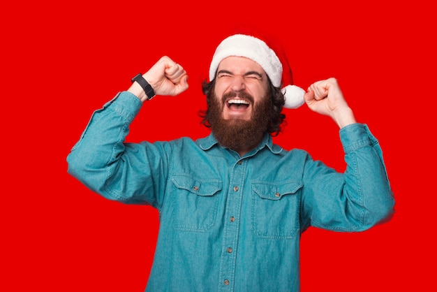 Finalmente as férias de inverno estão aqui, homem espantado com barba vestindo papai noel e comemorando com os punhos para cima