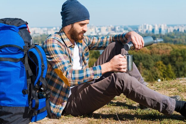 Finalmente posso relaxar. jovem bonito sentado perto da mochila e servindo chá em uma xícara