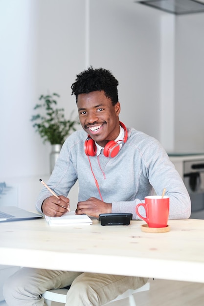 Financiador afro-americano contente com fones de ouvido e bloco de notas olhando para a câmera enquanto trabalha em casa
