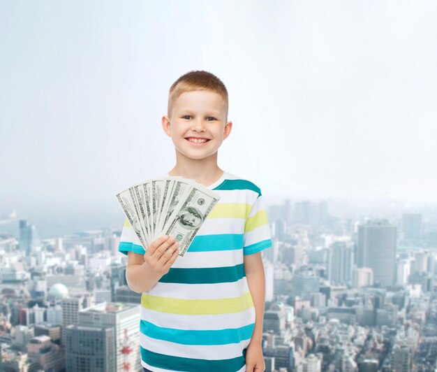 financiera, planificación, niñez y concepto - niño sonriente sosteniendo dinero en efectivo en dólares en su mano sobre los antecedentes de la ciudad