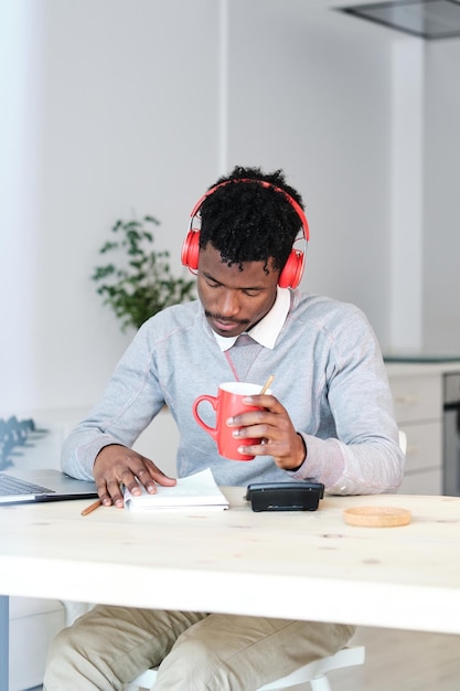 Financiero afroamericano en auriculares con una taza de bebida caliente trabajando en la mesa con computadora portátil y cuaderno en casa