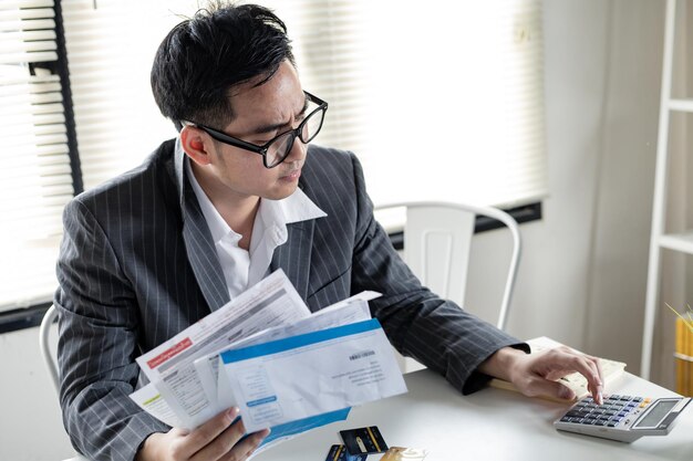 Finanzielles Problemkonzept. Man berechnet die Schulden mit einem Taschenrechner. Der junge asiatische Mann ist gestresst und überlegt durch Schulden von vielen Kreditkarten und Rechnungen.