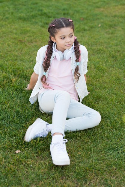 Fique feliz você mesmo Penteado de tranças de menina e fones de ouvido modernos aproveitem para relaxar Segredos para criar uma criança feliz Menina linda criança fundo de grama verde Criança feliz emocional saudável relaxando ao ar livre