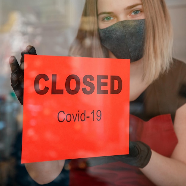 Foto firmar cerrado covid 19 lockdown en la puerta de entrada de la tienda como nuevo cierre normal. mujer en guantes de máscara médica protectora cuelga el cartel de cerrado en la ventana del restaurante cafetería. cultivo de lockdown square.