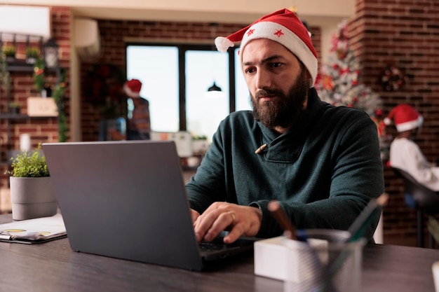 Firmenangestellter mit Weihnachtsmütze, der im festlichen Büro, Weihnachtsbaumbeleuchtung und Dekorationen am Laptop arbeitet. Verwendung des Computers am Arbeitsplatz mit saisonalen Feiertagsornamenten und Weihnachtsdekor.