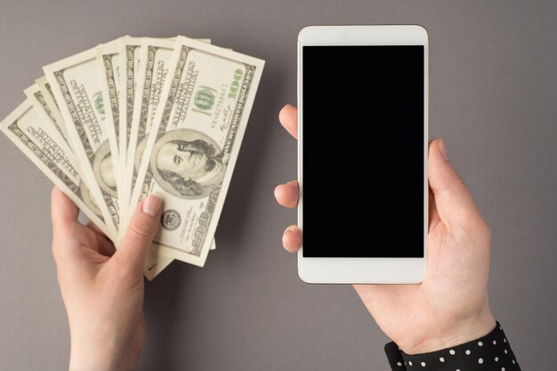 First-Person-Draufsichtfoto von Frauenhänden, die Smartphone und Fan von Geldhundert-Dollar-Banknoten auf isoliertem grauem Hintergrund mit Copyspace halten