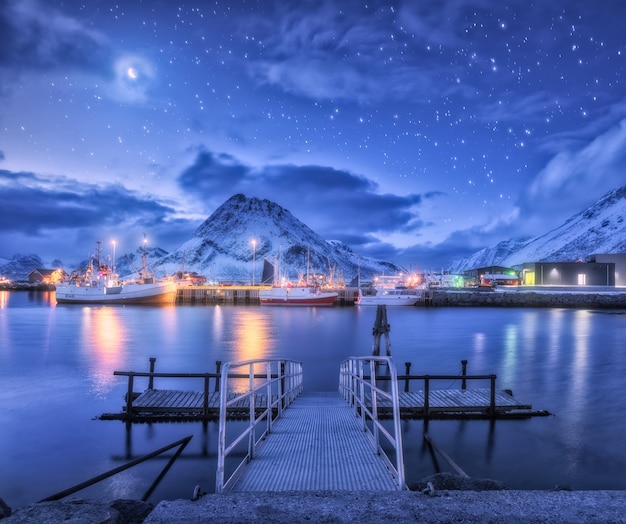 Fischerboote nahe Pier auf dem Meer gegen schneebedeckte Berge und Sternenhimmel mit Mond bei Nacht in den Lofoten-Inseln, Norwegen.