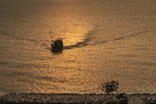 Fischerboote segeln im Meer und näherten sich dem Ufer im Abendsonnenuntergang mit goldener Sonne, die hell strahlte, warme und romantische Landschaft, natürliches Seereisekonzept