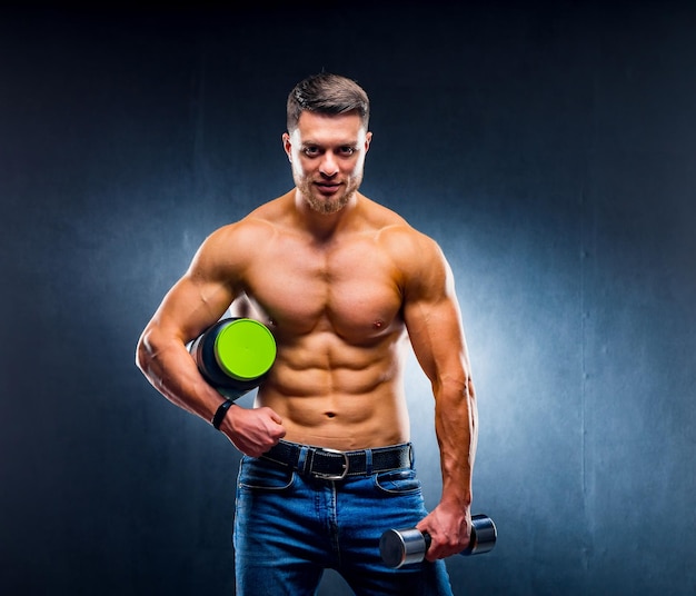 Fisiculturista atleta masculino segura um pote de nutrição esportiva e haltere nas mãos Posando para as poses da câmera sobre fundo azul