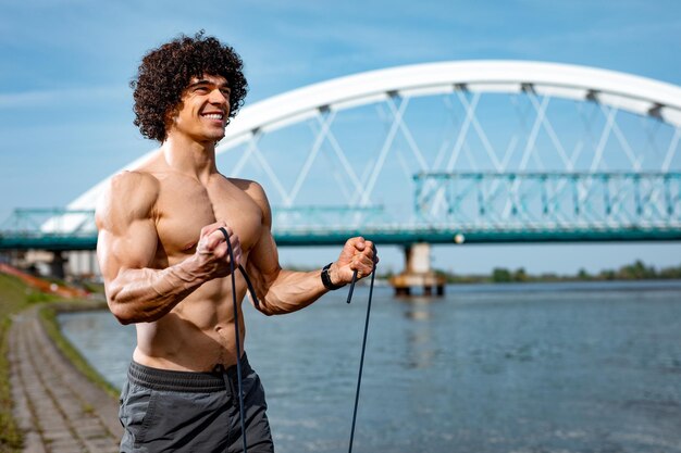 Fisiculturista com torso nu, está fazendo treinamento de corpo em forma forte com elástico de borracha, perto do rio.