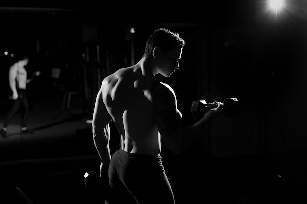 Fisiculturista de cara atlético bonito poder fazendo exercícios com halteres. Corpo musculoso de aptidão em fundo escuro. Preto e branco
