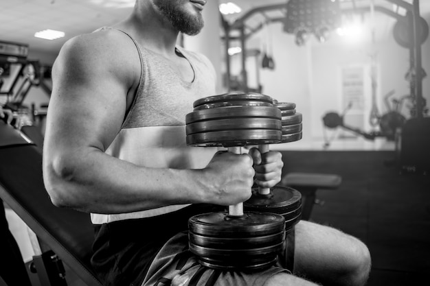 Fisiculturista de homem forte está sentado com halteres pesados no ginásio. corpo musculoso de um esportista no centro esportivo. foto em preto e branco