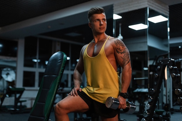 Foto fisiculturista jovem desportivo bonito com corpo musculoso e tatuagem em regata amarela treinando com halteres no ginásio