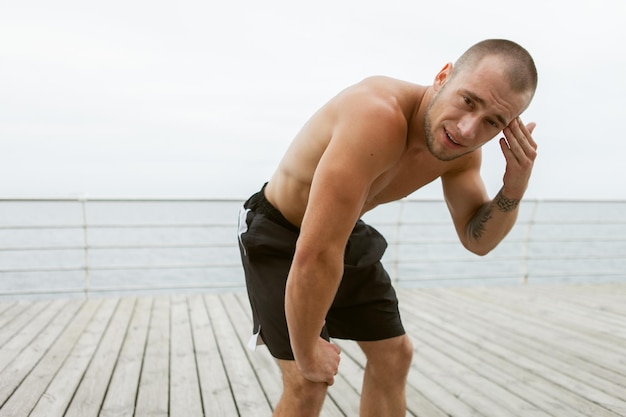 Fisiculturista masculino cansado com um torso nu enxuga o suor da testa após um treino intenso na praia