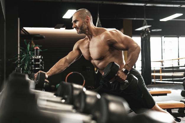 Fisiculturista massivo fazendo remada com halteres com um braço durante seu treino em uma academia