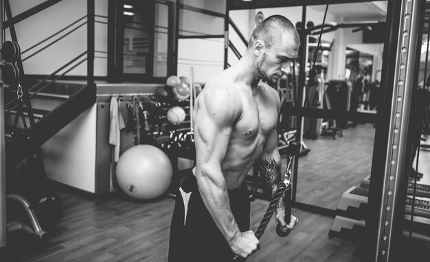 Fisiculturista musculoso homem bonito com torso nu treina tríceps em uma máquina de exercícios com cabo cruzado em uma academia moderna