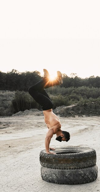 Fisiculturista profissional masculina fazendo exercícios para braços em pneus ao ar livre. Jovem com máscara preta treinando regularmente para manter a forma do corpo musculoso.