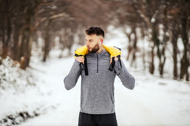 Fisiculturista segurando pesos em pé na floresta em dia de inverno nevado. Musculação, fitness de inverno