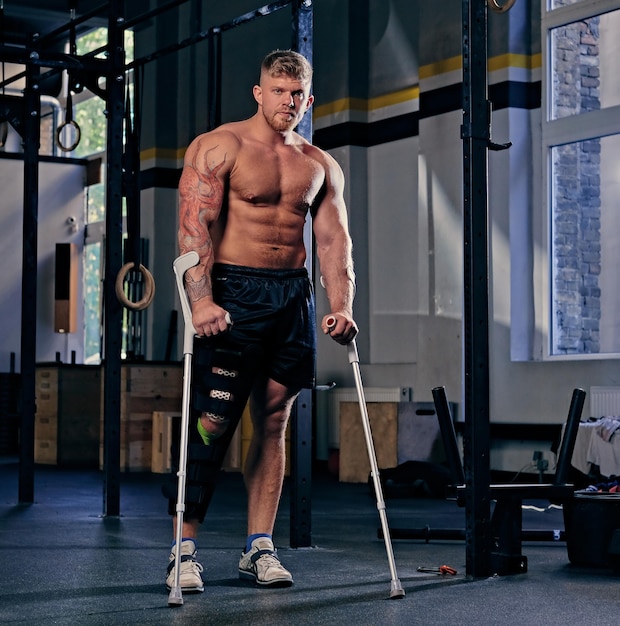 Fisiculturista sem camisa em muletas perto de cross fit stand em uma academia.