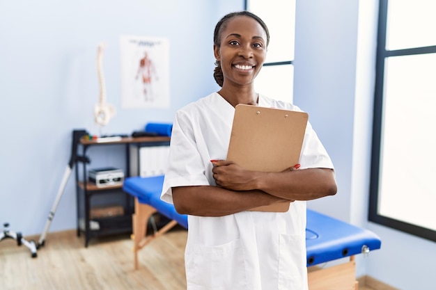 Fisioterapeuta afroamericana sonriendo confiada sosteniendo un informe médico en la clínica de rehabilitación